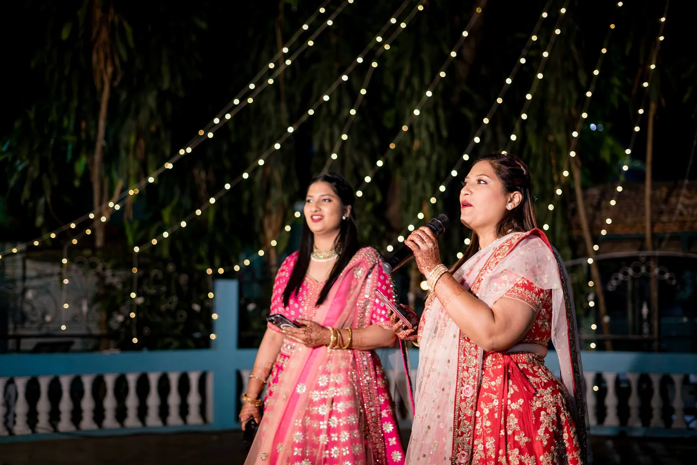 Two women in pink and red lehengas look up and laugh on the terrace with the blue railing behind them