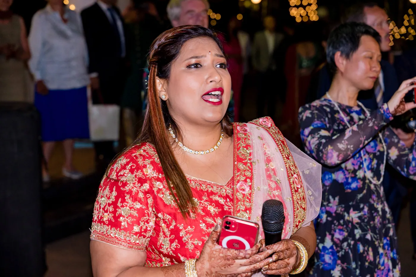 A woman in a red embroidered lehenga holds a microphone and sings along with an open mouth