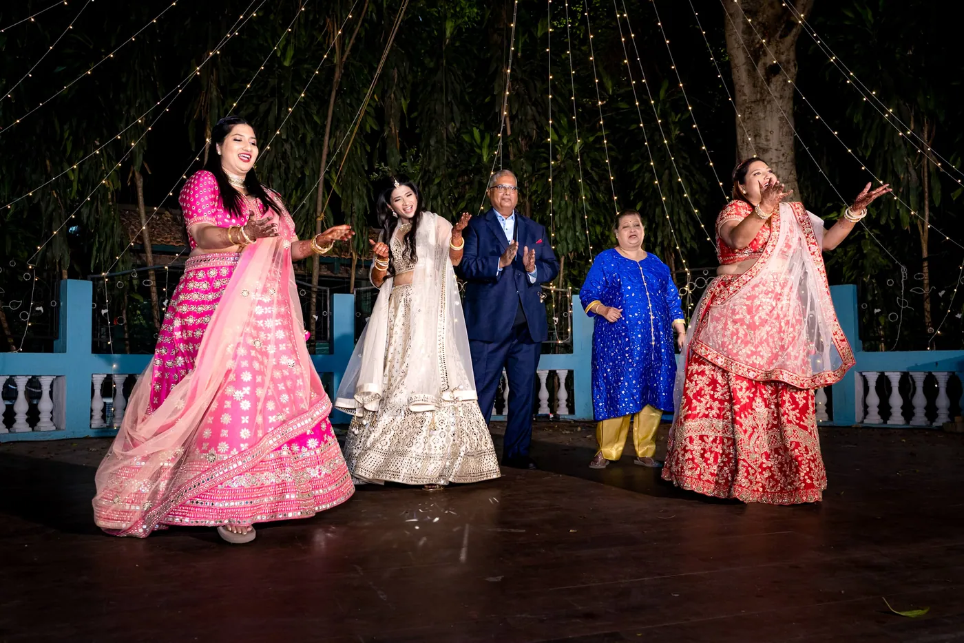 Medha dances between two of her sisters in pink and red lehengas on the dark wooden floor