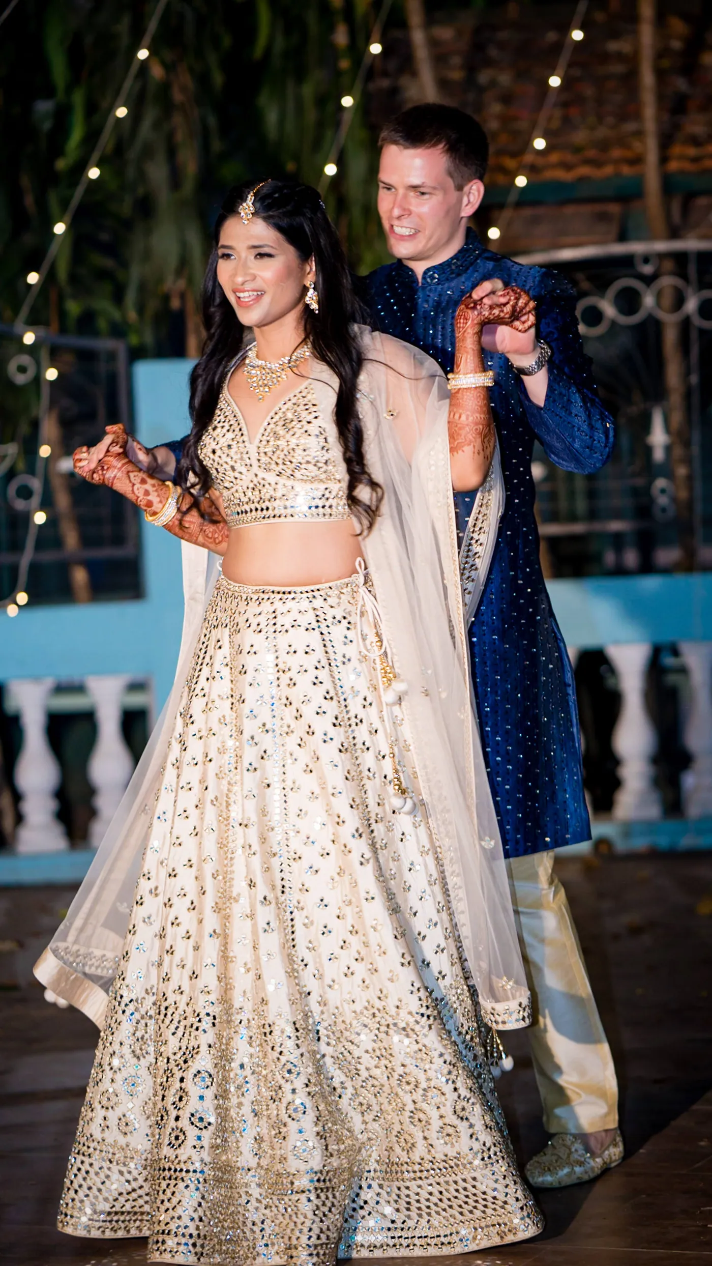 Medha in her cream lehenga dances forward with a smile as Stefan moves behind her