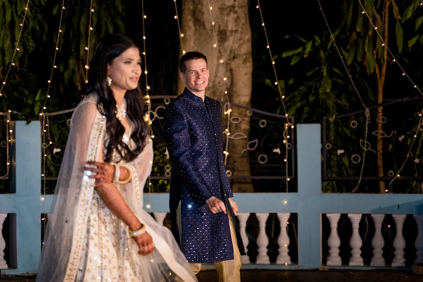 Medha smiles to the side beside Stefan on the open dance floor with the blue balustrade and fairy lights behind