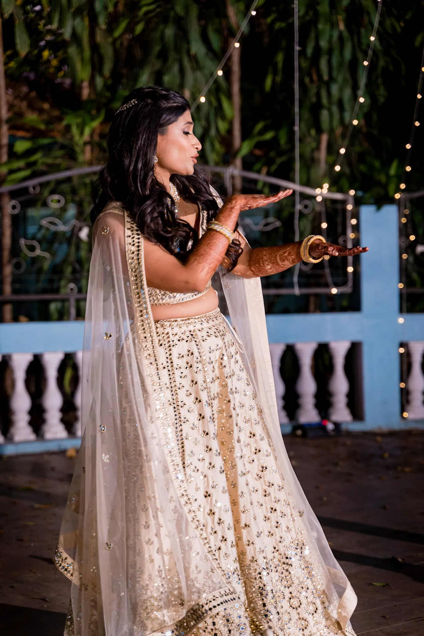 Medha in a cream lehenga sings or sways with her arm stretched out near the blue railing