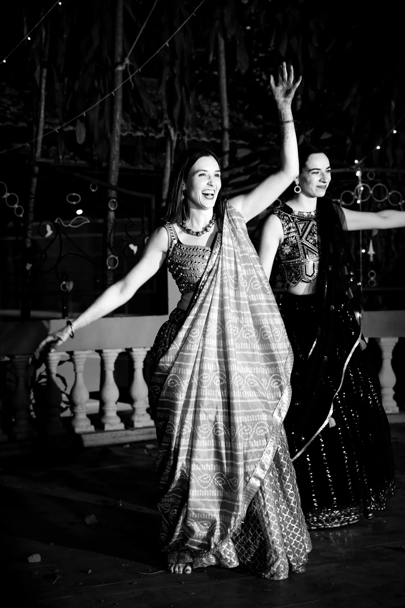 In black and white, two women in saris dance with their arms raised high
