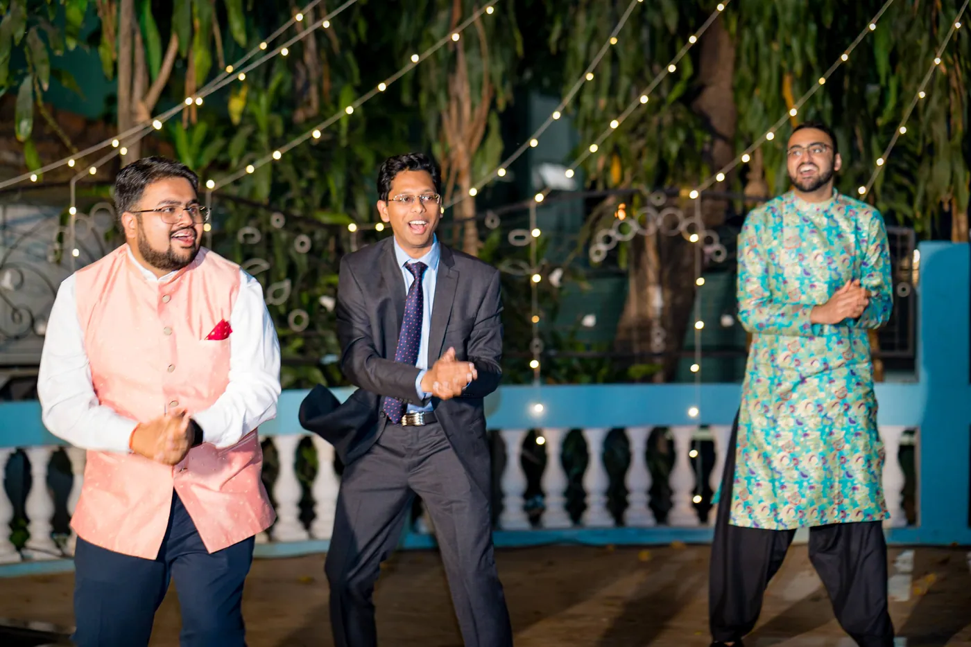 Three men in a pink waistcoat, a navy suit and a teal kurta stand grinning on the floor