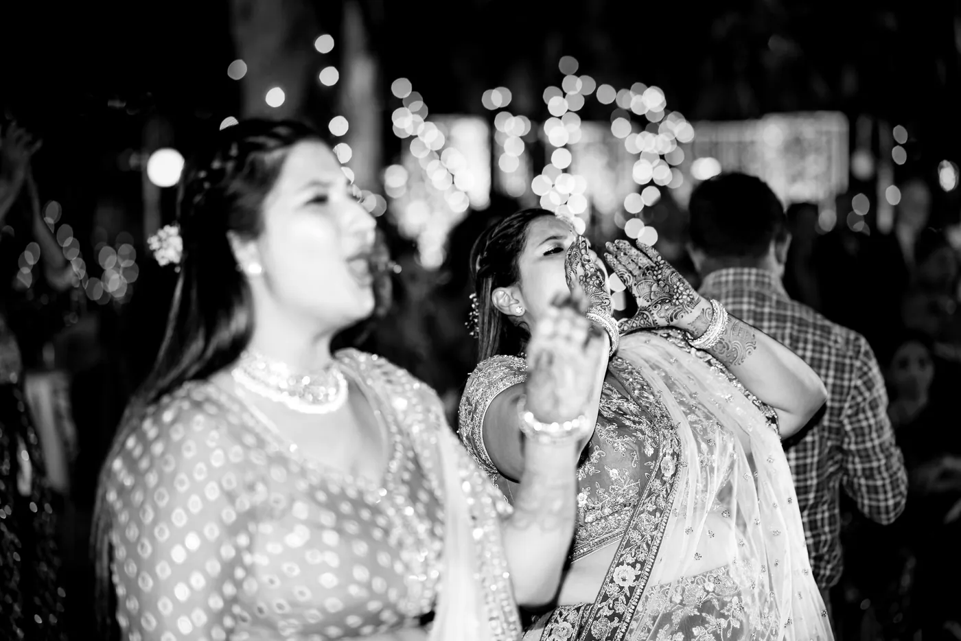 In black and white, two women in sparkling lehengas laugh together with open mouths