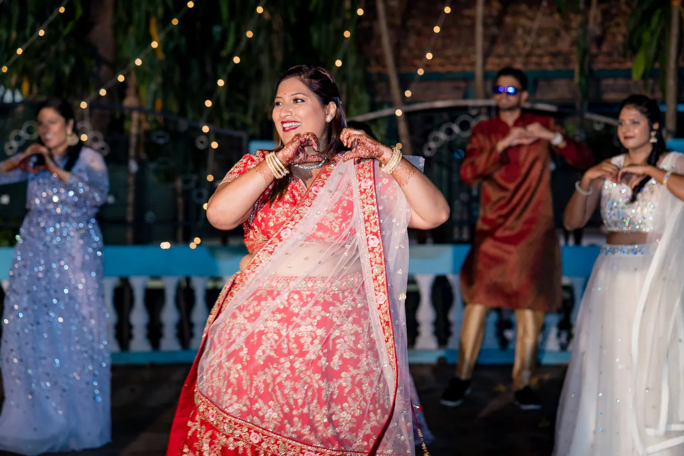 A woman in a coral lehenga dances with her arms in front while others dance in pale dresses