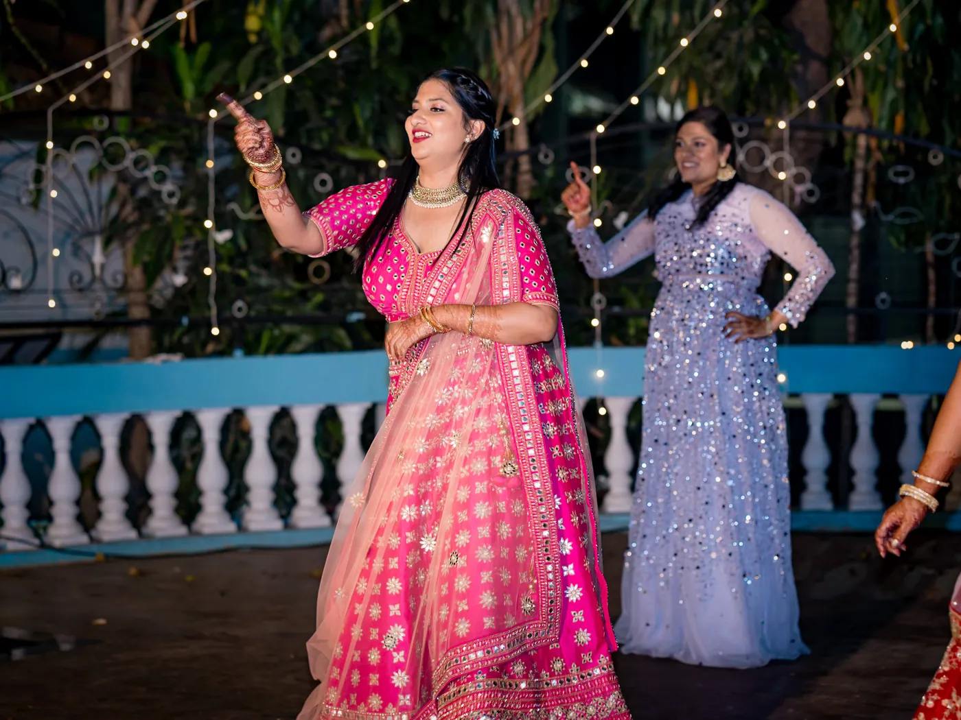 A woman in a hot pink lehenga and a woman in lilac sequins dance together by the blue railing