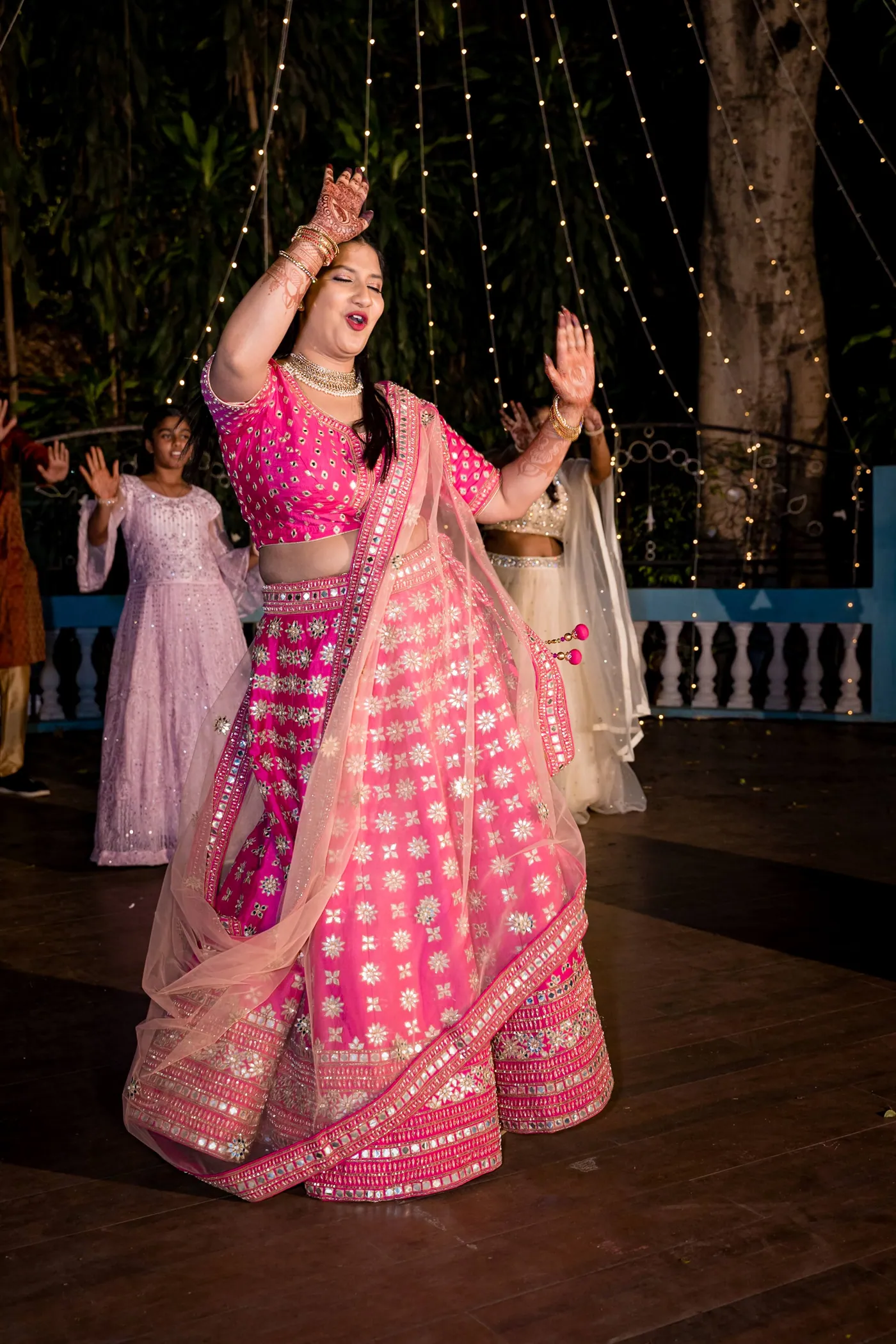 A woman in a pink lehenga lifts one arm mid dance with the blue railing behind her