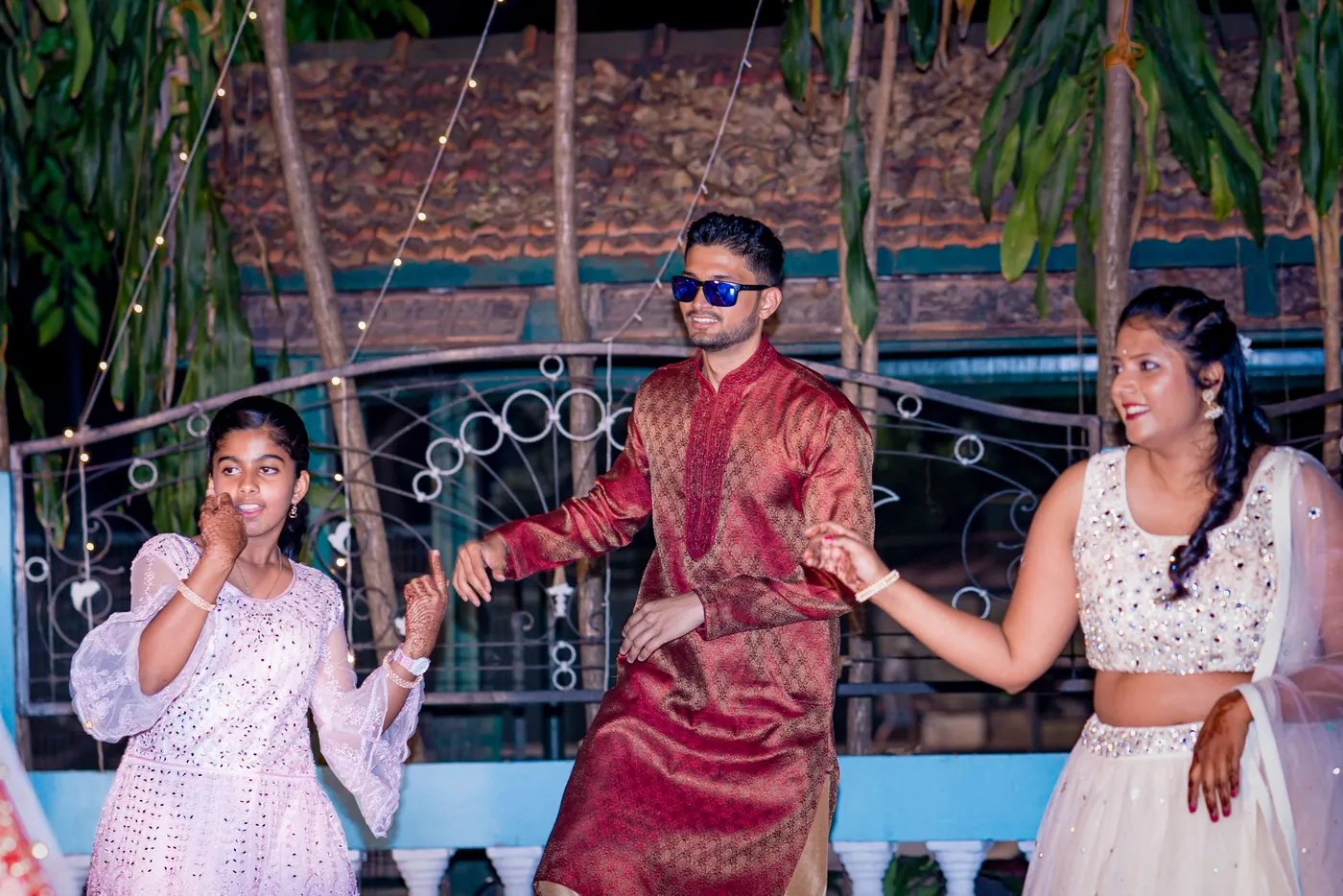 A man in a red kurta and sunglasses dances between two women in pale outfits