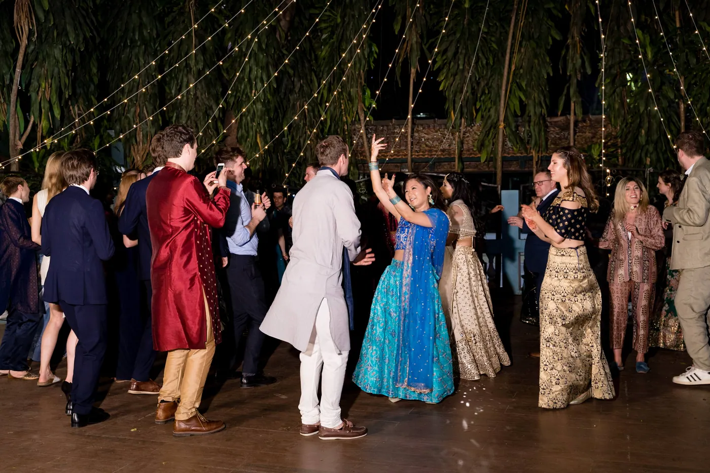 A crowd of guests in saris, suits and kurtas dances across a wide wooden floor