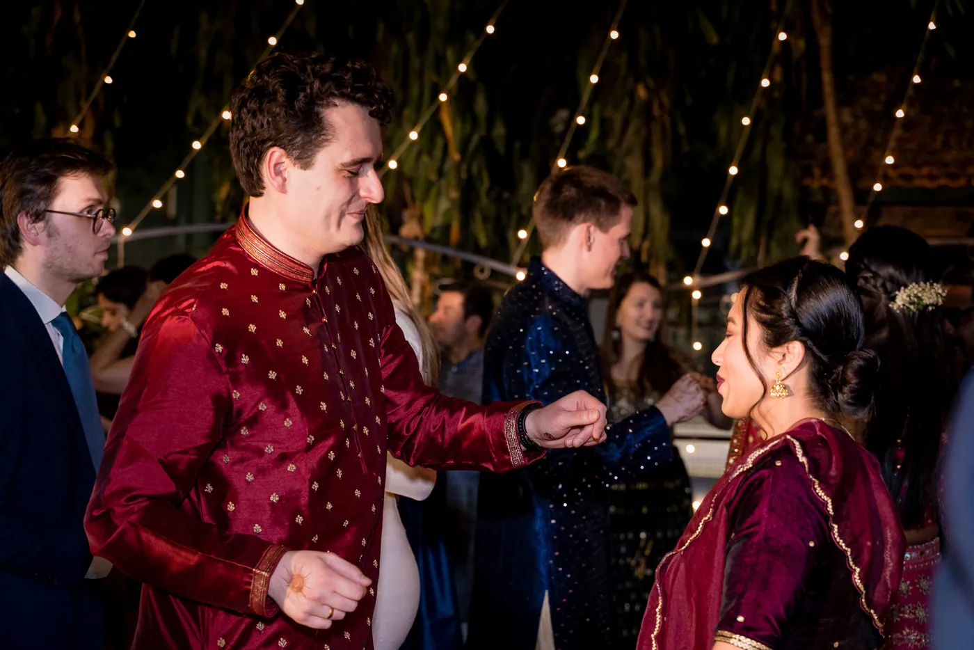 A man in a maroon kurta and a woman in a maroon dress dance while others in navy gather around