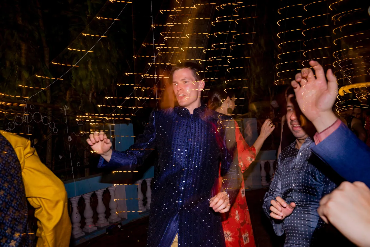 Stefan raises one arm and dances in a navy sherwani as friends swirl around him