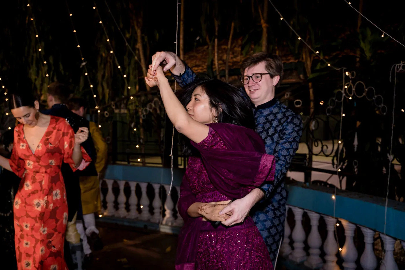 A woman in a magenta dress dances with a man in glasses and a navy sherwani behind her