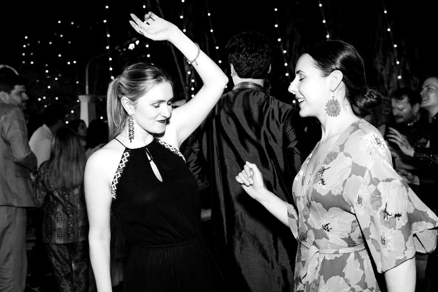 In black and white, a woman in a halter dress dances with an arm overhead beside another woman laughing
