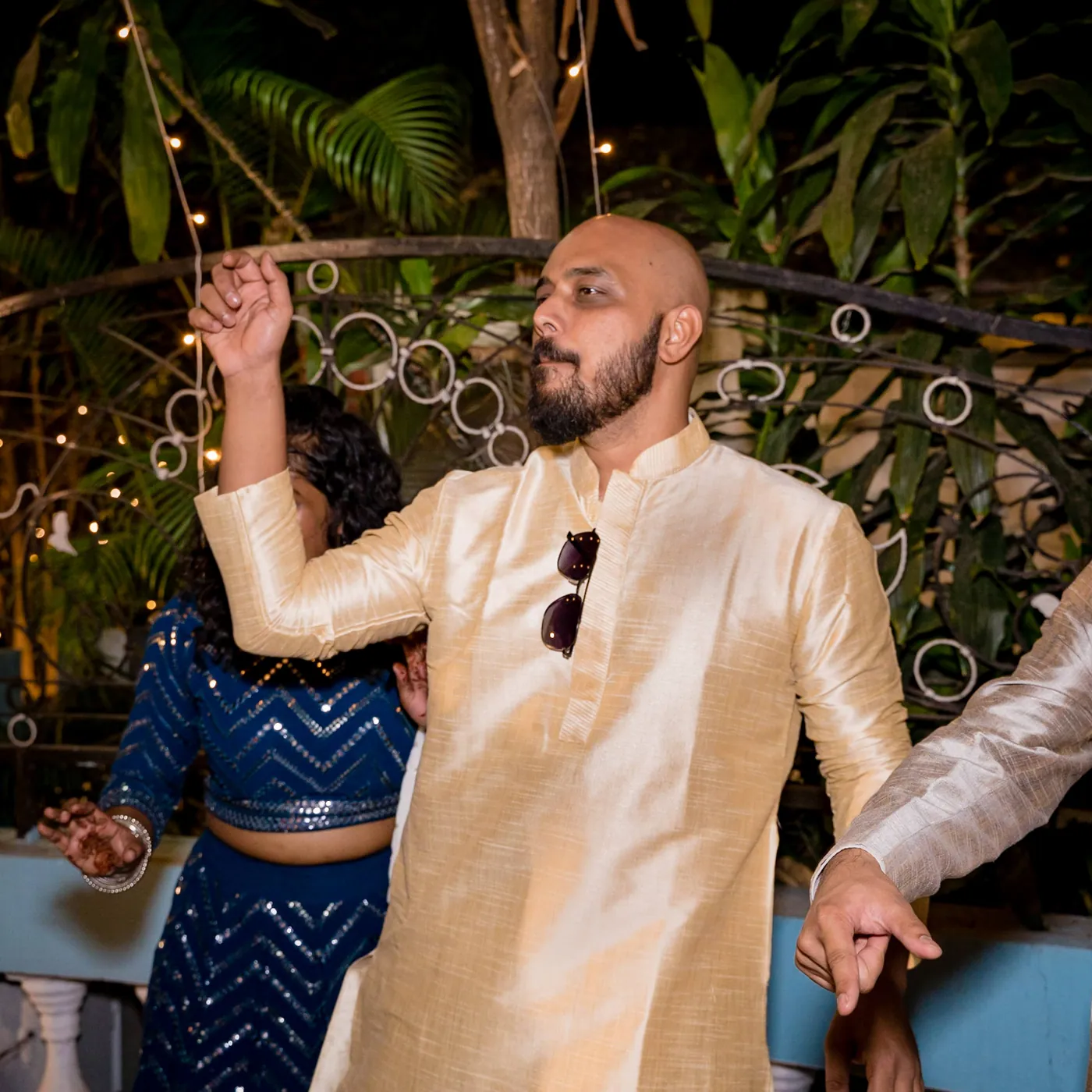 A bearded man in a gold kurta dances with one arm up and sunglasses hooked on his collar
