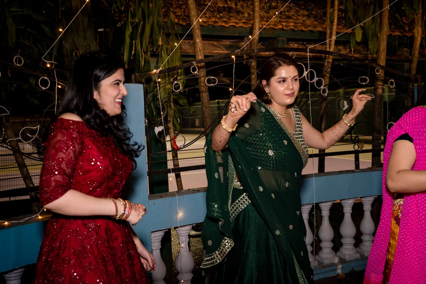 A woman in a red lehenga and a woman in a green sari laugh and dance together