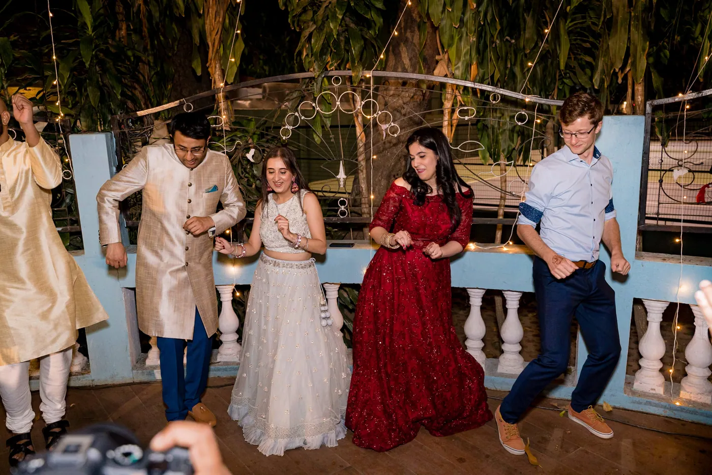 A row of guests in lehengas and kurtas dances along the terrace while a photographer's hand shows in the corner