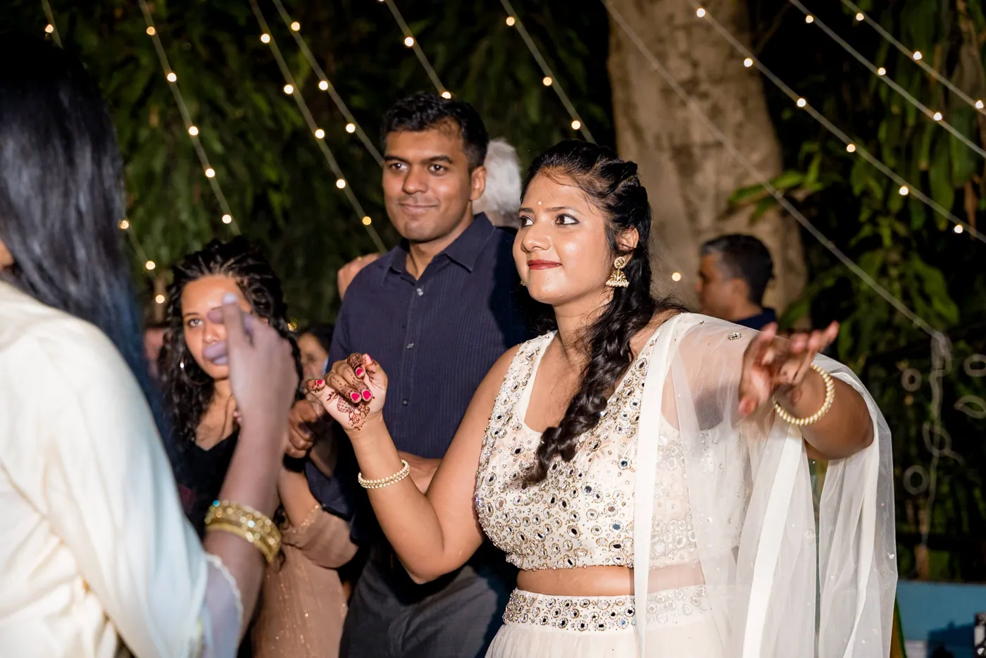 A woman in a white lehenga dances with a smile among guests under strings of lights