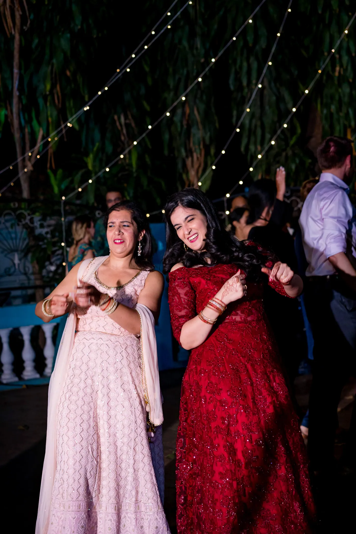 Two women in a pastel lehenga and a red lehenga dance together