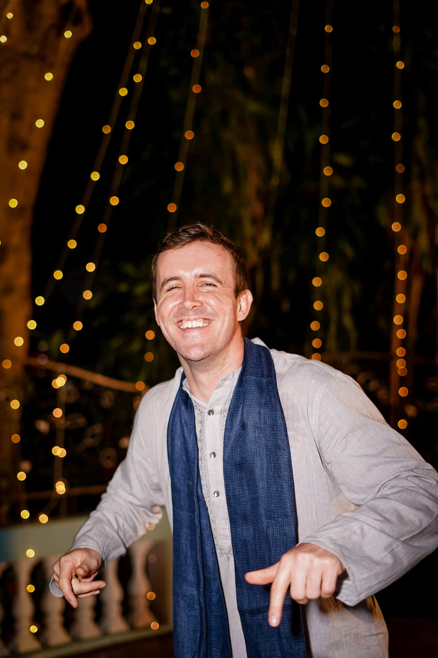A man in a pale kurta and a navy shawl dances mid laugh with arms lowered