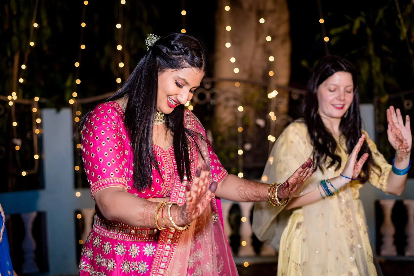 A woman in a pink lehenga laughs and dances with a woman in a cream outfit