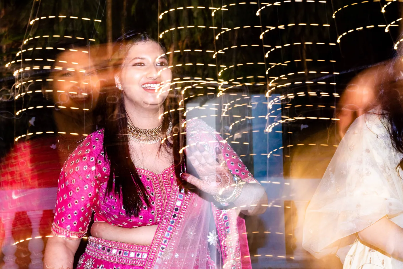 A motion blur wraps around a woman in a pink lehenga laughing as lights streak behind her