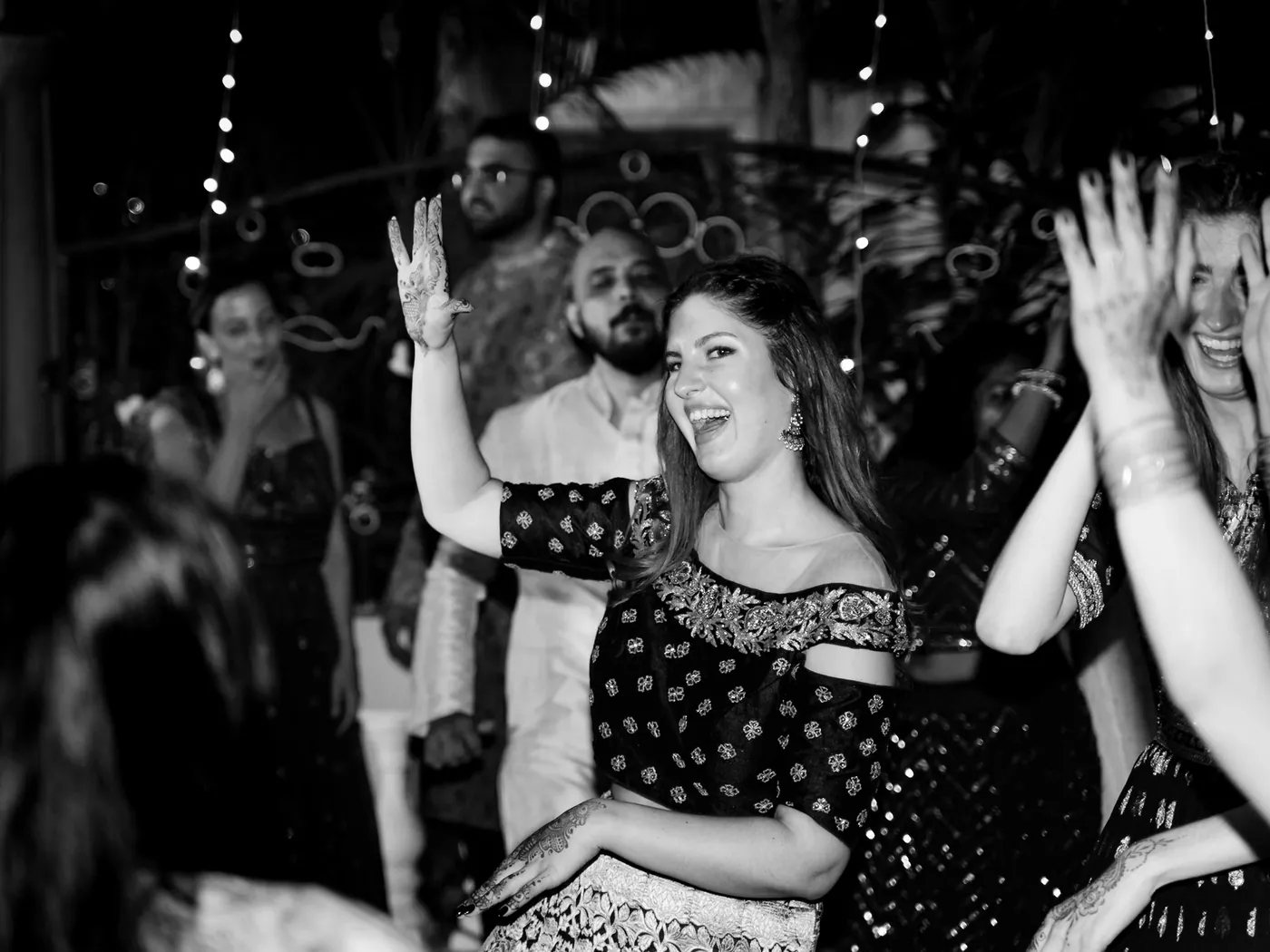 In black and white, a woman in a dark dress laughs among dancing guests with arms in the air