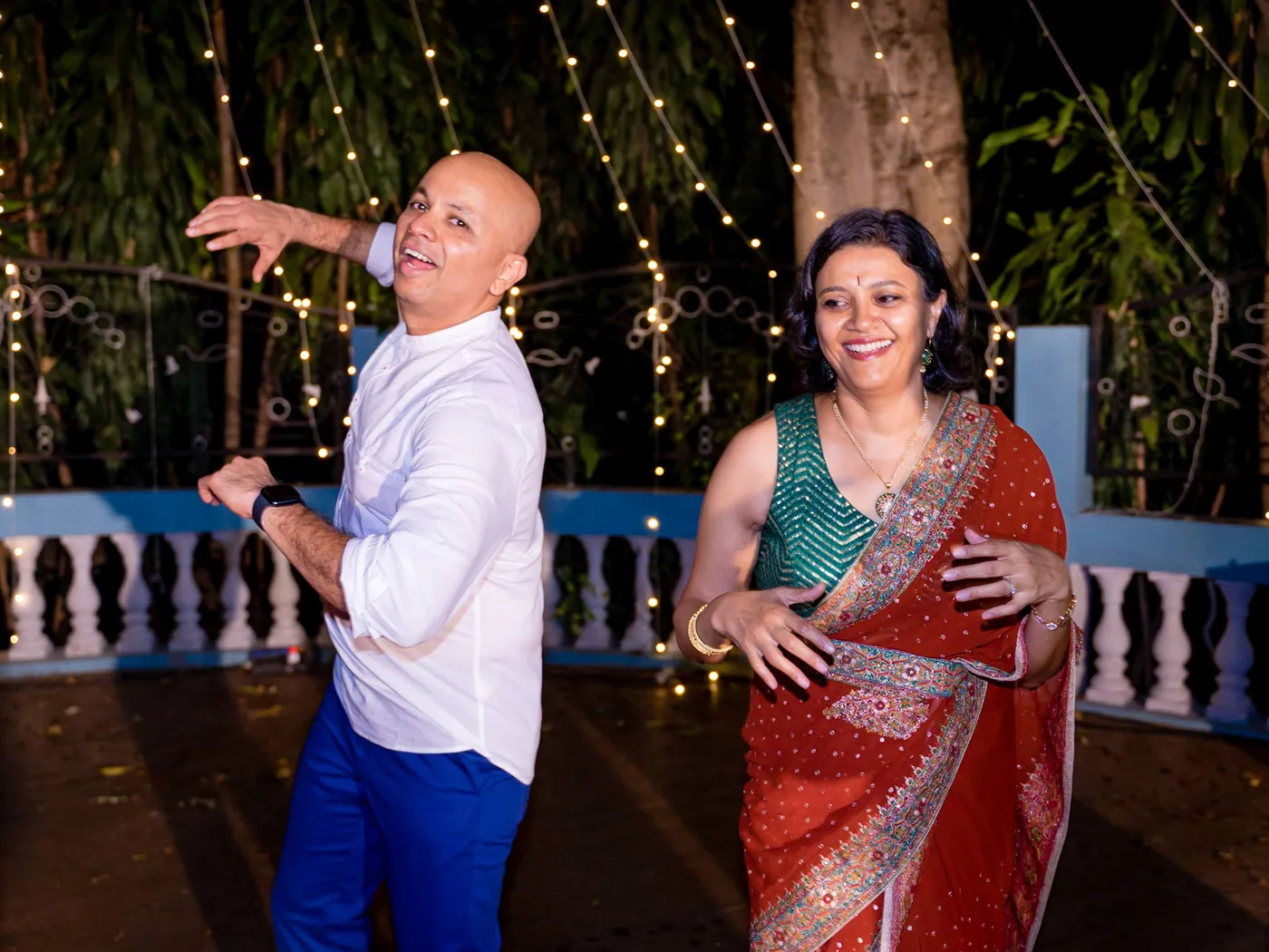 A bald man in a white shirt and a woman in a red sari dance side by side