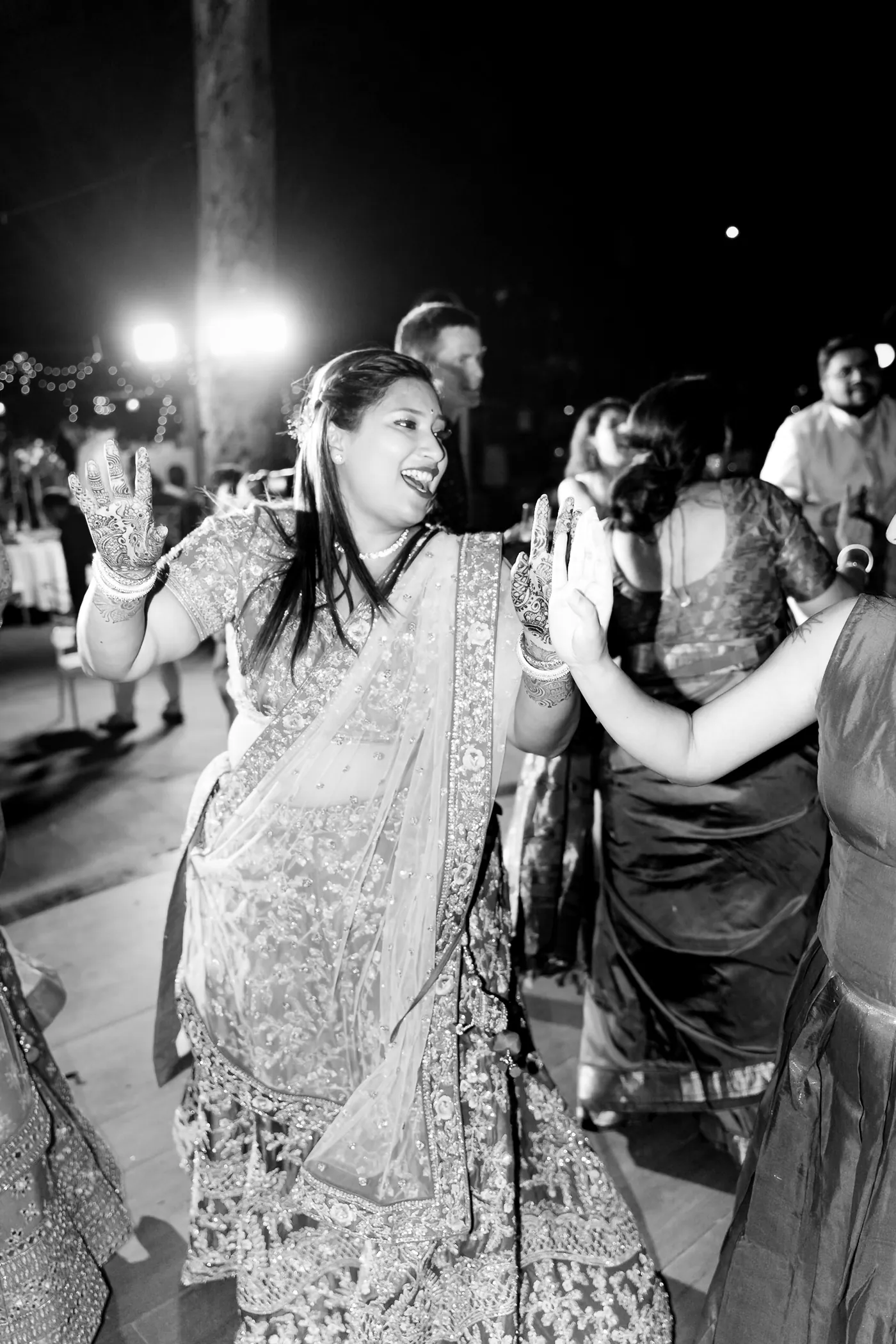 In black and white, women in lehengas dance and laugh near a bright light