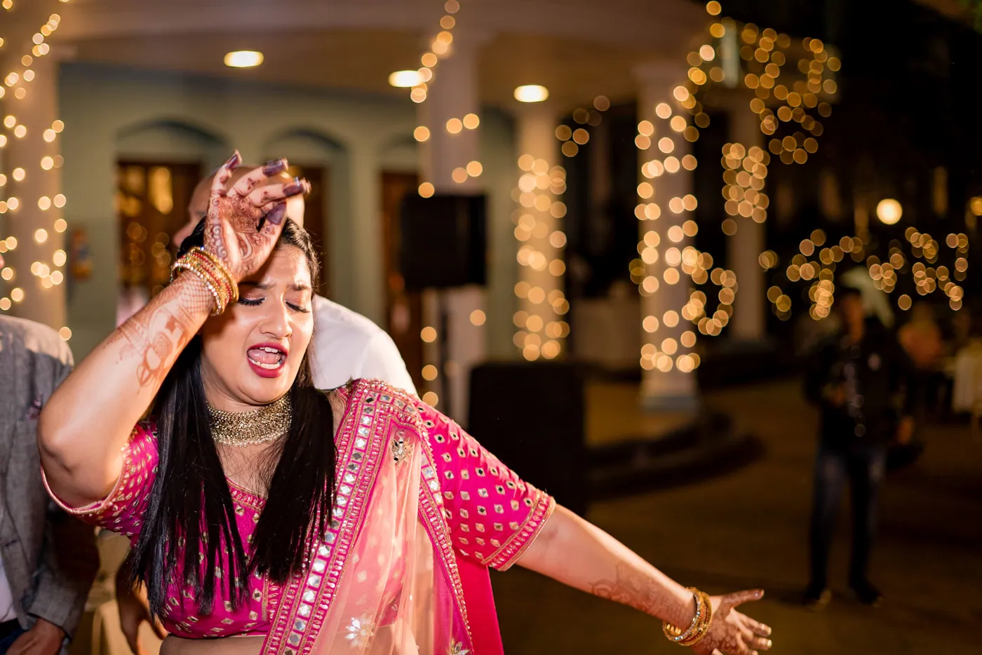 A woman in a pink and gold lehenga dances with an arm stretched wide and a big grin