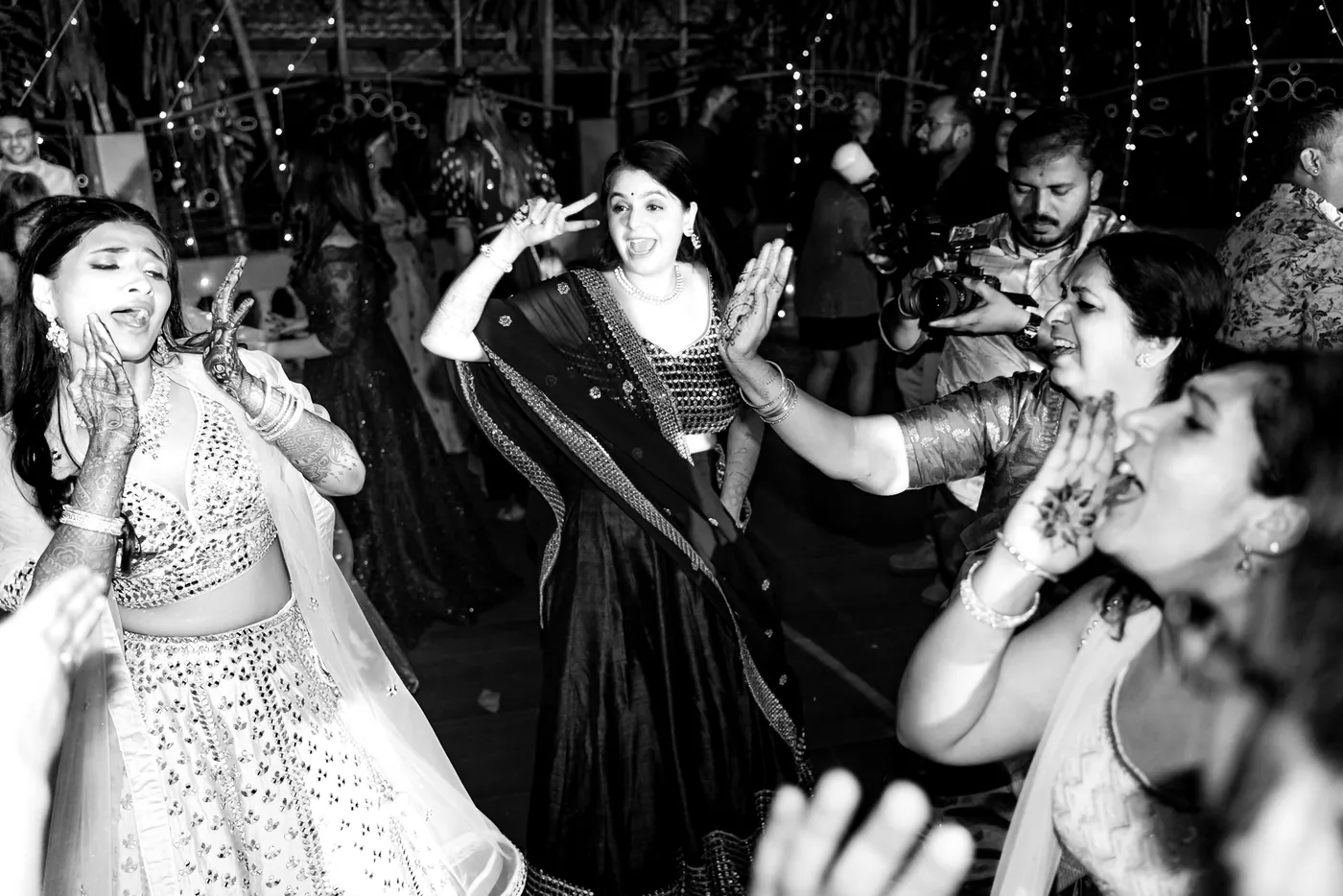 In black and white, a woman in a light lehenga dances with her hand to her face among a crowd