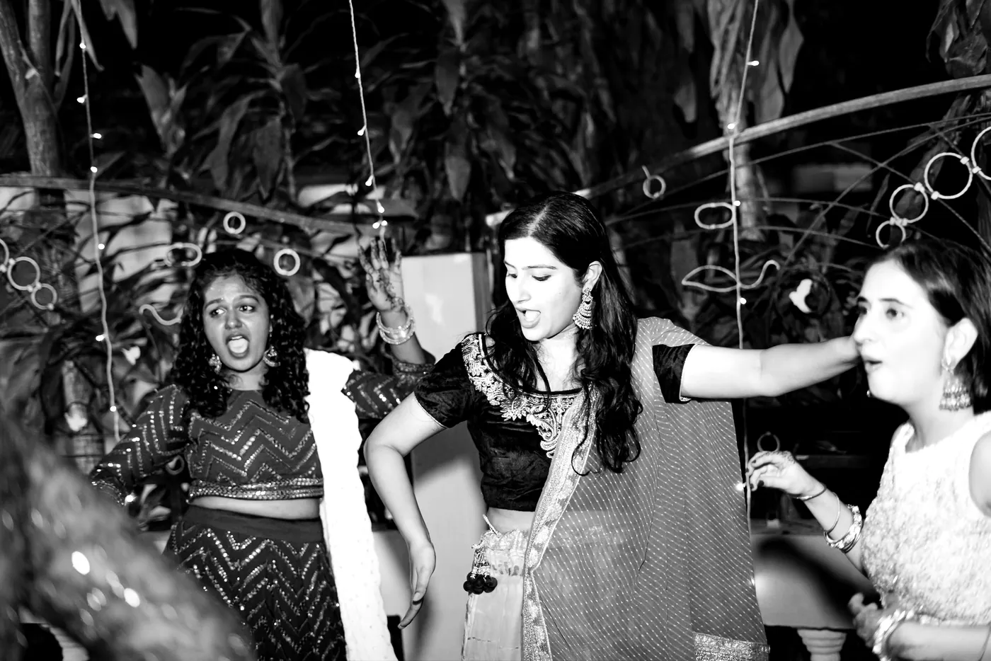 In black and white, women in dark tops and light lehengas cheer and dance together