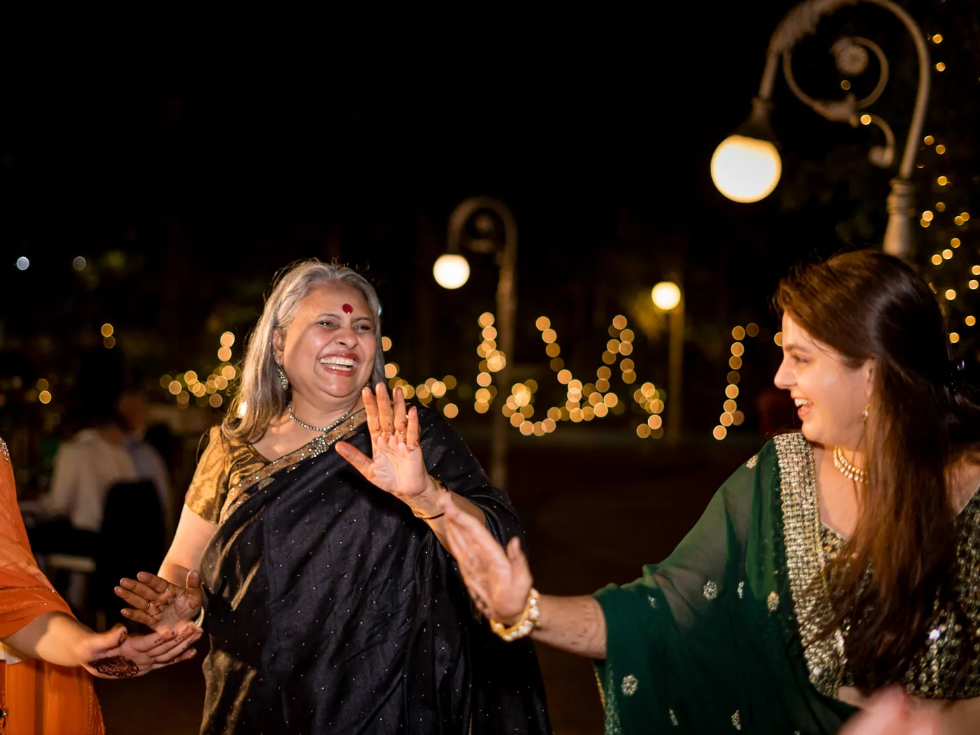 A woman in a navy sari dances with a blonde woman in a green dress, both laughing