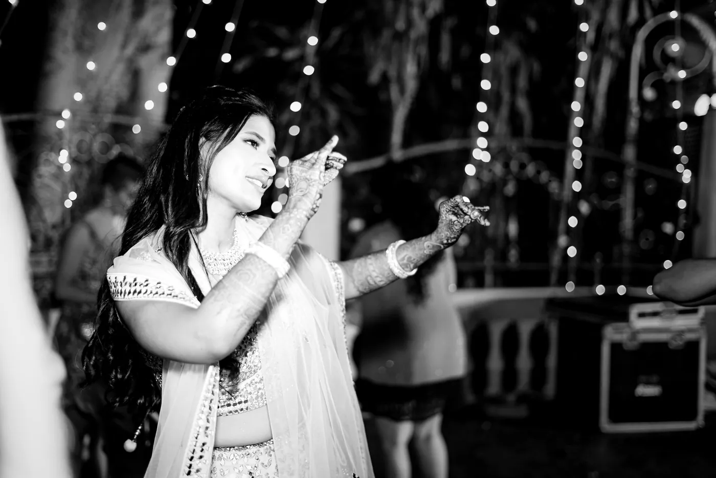 In black and white, Medha in a lehenga sings or points with one arm raised in front of the dancers