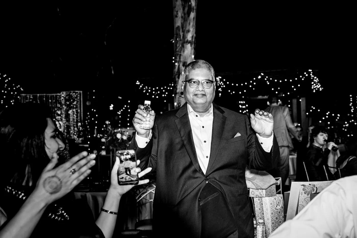 In black and white, a man in a dark suit stands calmly among guests holding up a phone