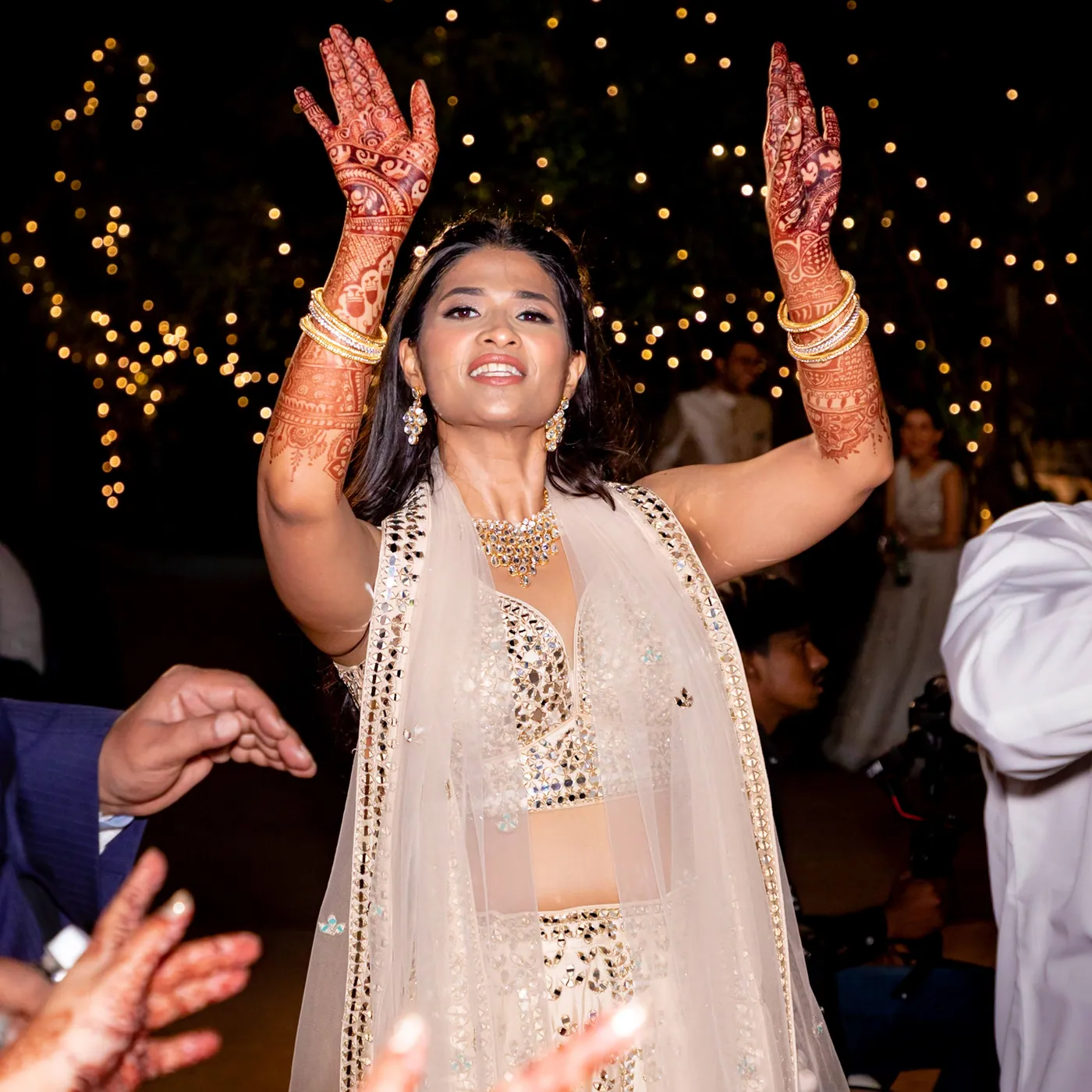 Medha throws both hands in the air in her cream lehenga with henna on her arms and a wide smile