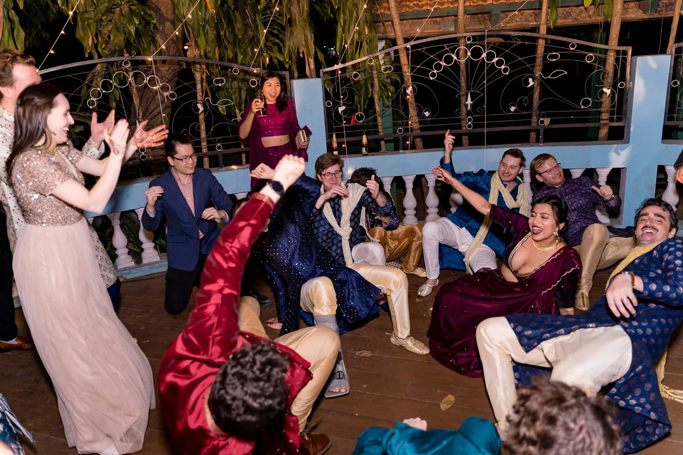 A group of men in sherwanis and kurtas crouch and fall to the floor laughing while women dance behind them