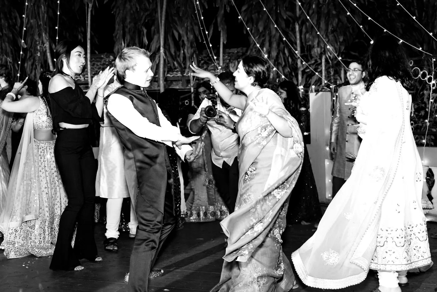 In black and white, a man in a waistcoat and women in lehengas dance in a crowd