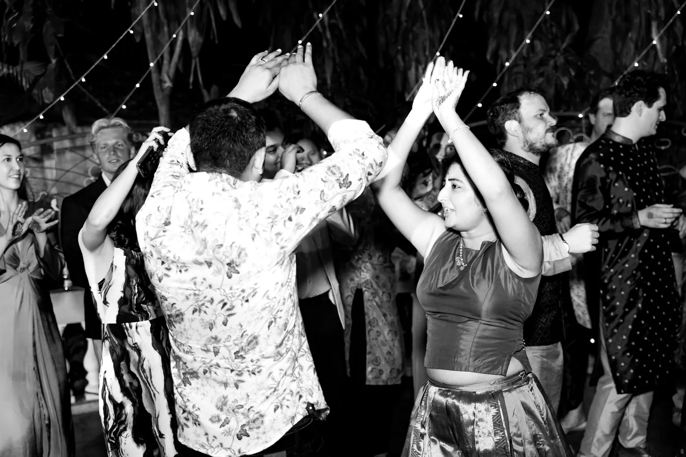 In black and white, men and women in shirts and dresses dance with their arms above their heads