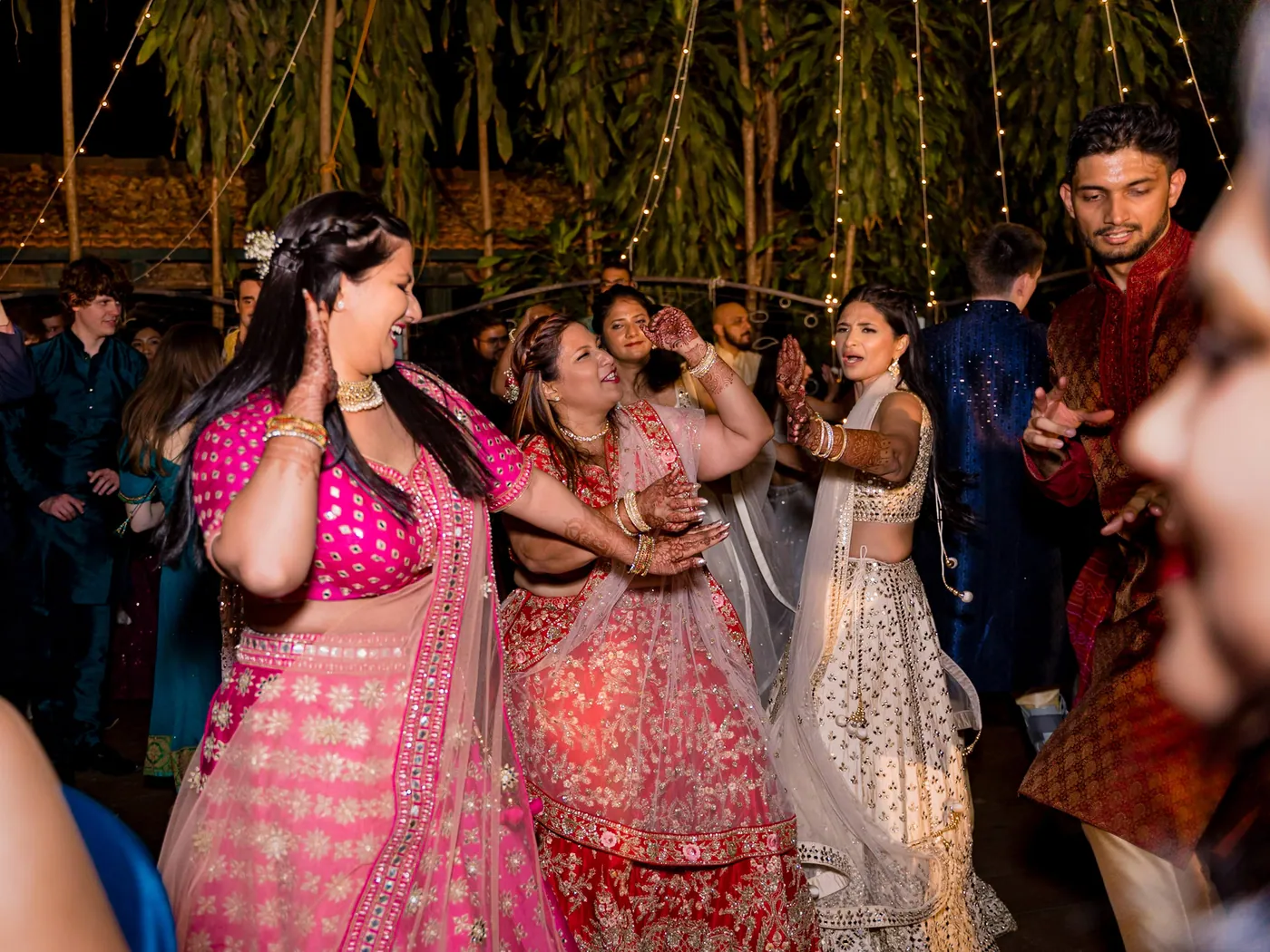 Women in pink and red lehengas dance with big smiles among the crowd