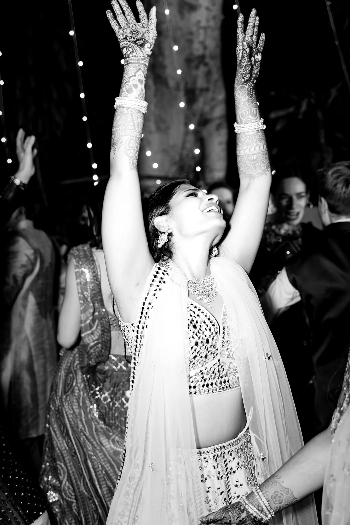 In black and white, a woman in a cream lehenga throws both arms up high