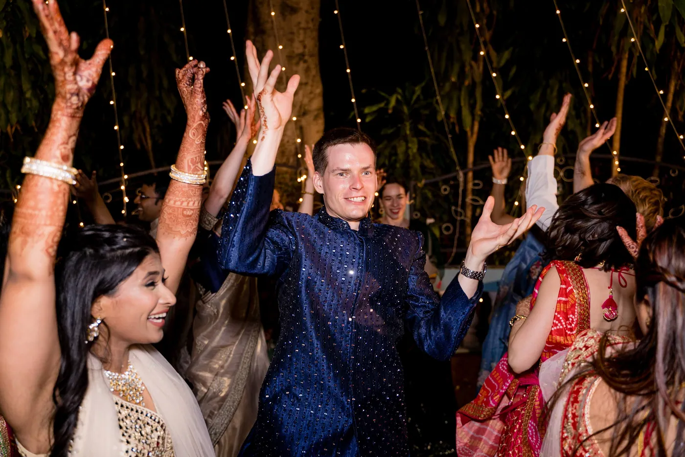 Stefan in navy raises his hands as Medha, with hennaed arms in the air, dances next to him