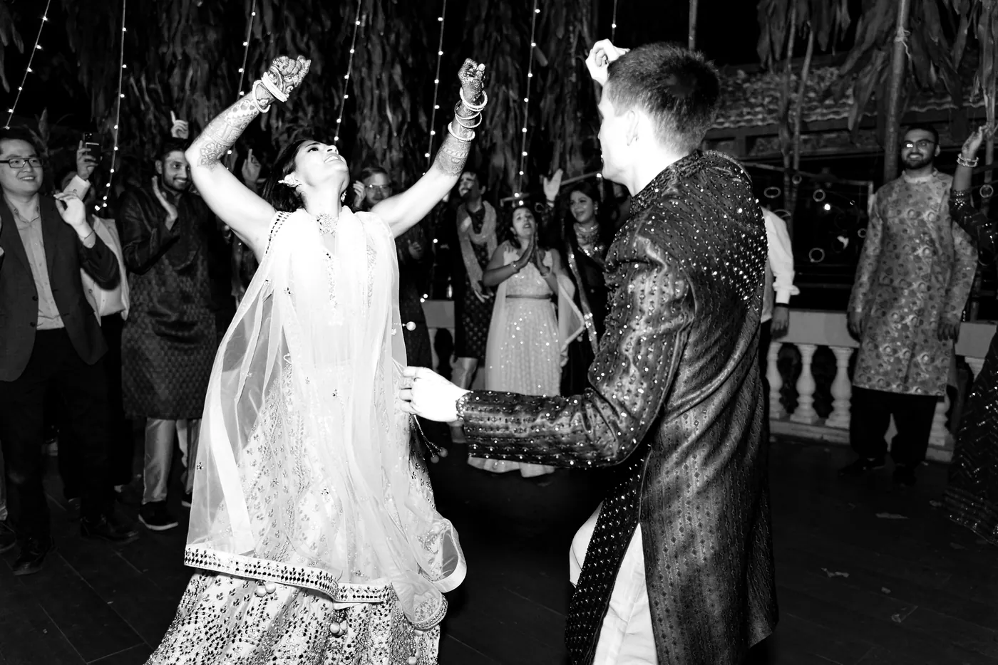 In black and white, Medha raises her arms while Stefan dances in a patterned sherwani next to her