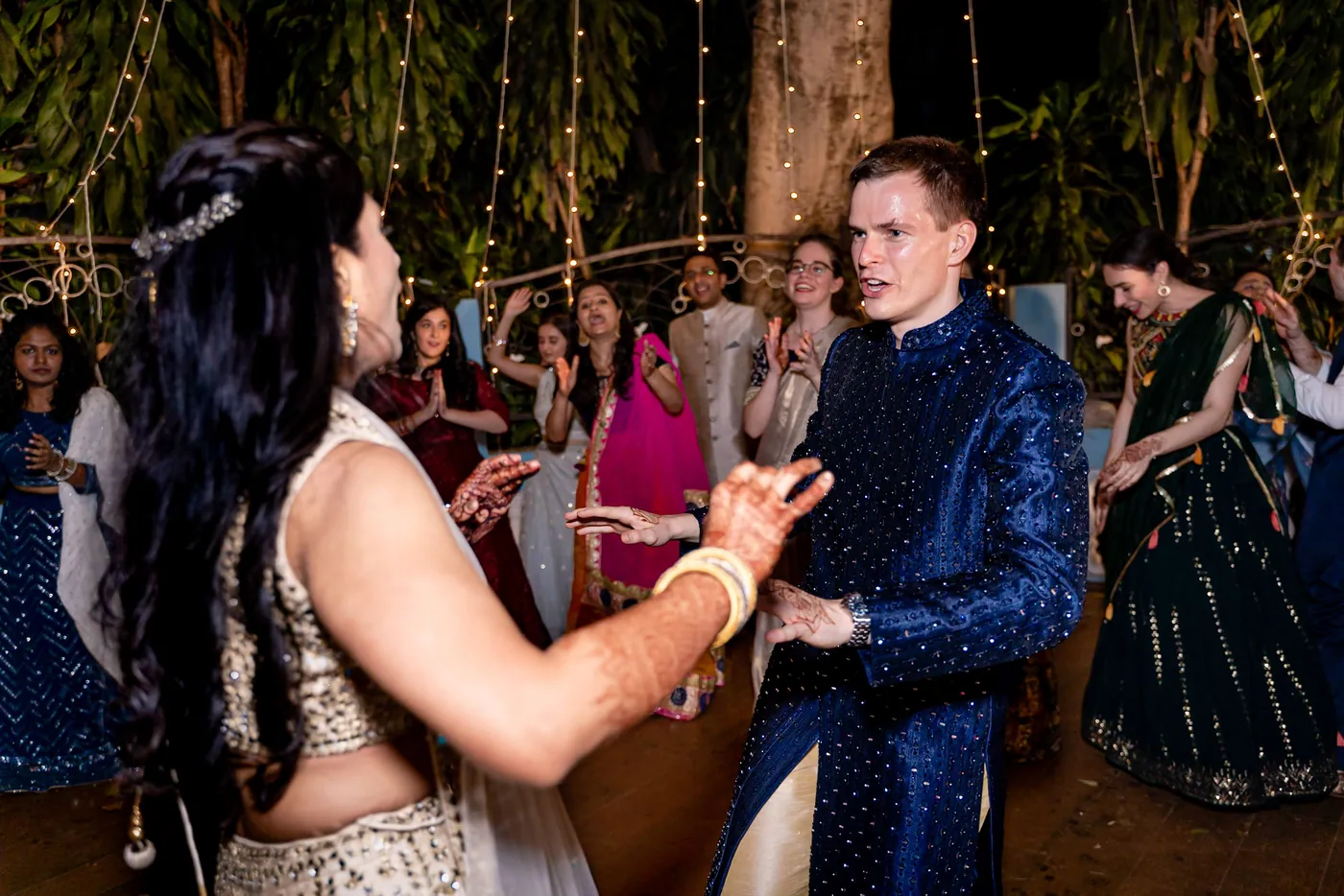 Medha with a hair ornament dances toward Stefan in his navy sherwani, both focused on each other