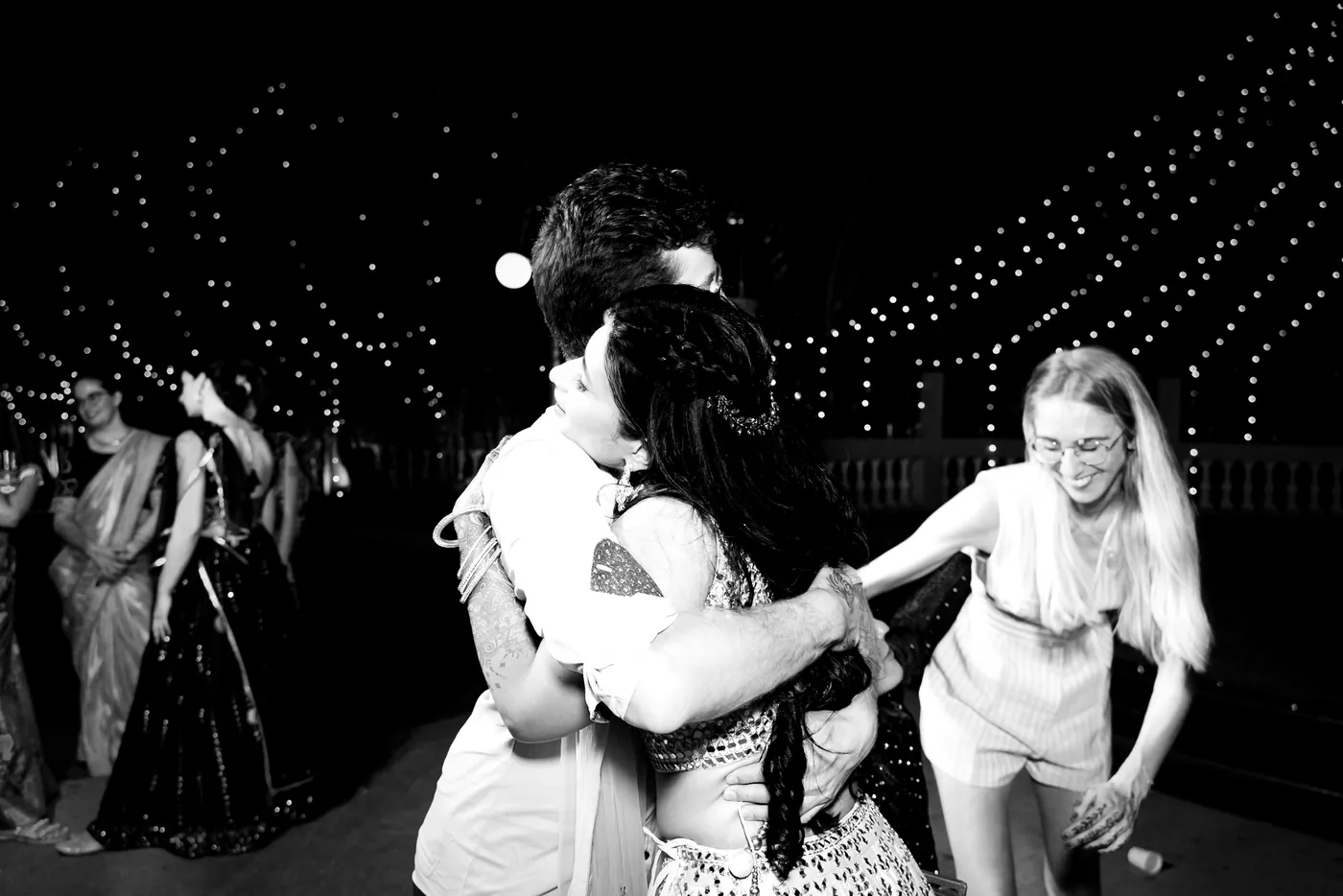 In black and white, a blonde woman in a white romper hugs Medha as others dance behind them