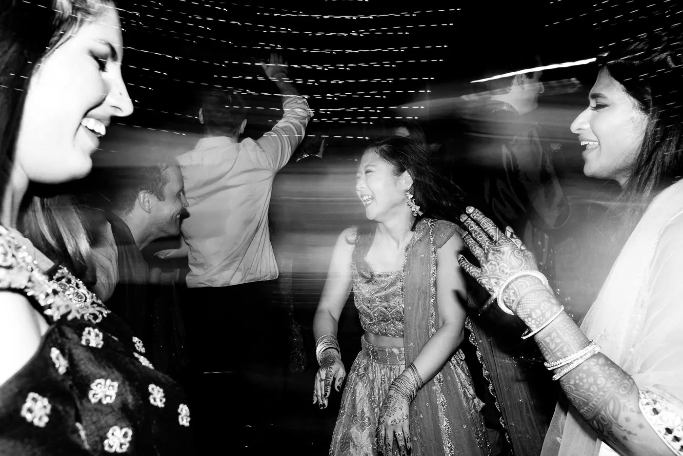 Motion blur streaks across the floor as women laugh and dance with henna hands raised