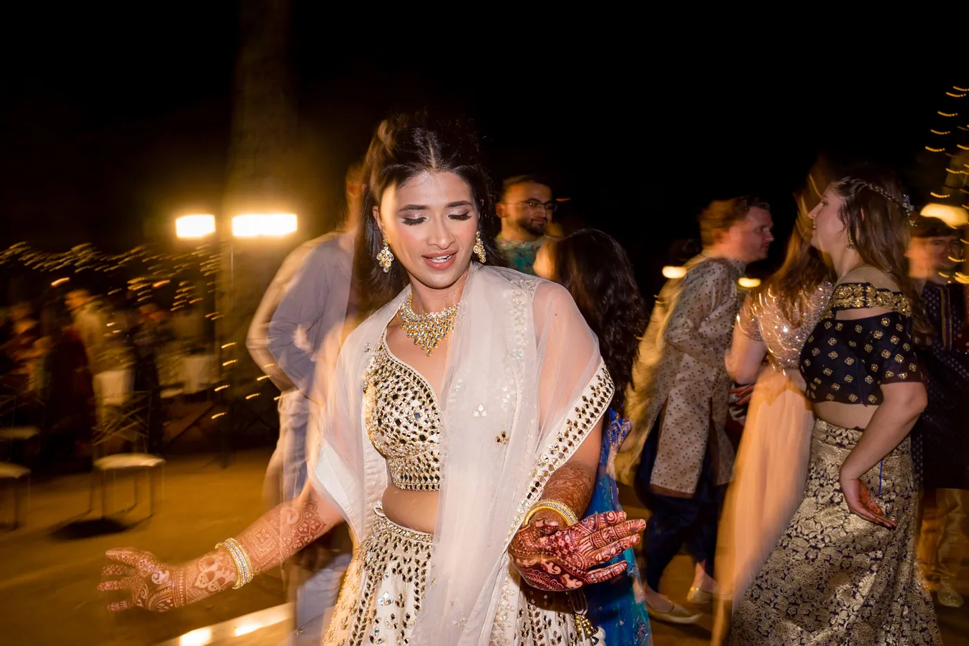 Medha laughs mid dance in her cream lehenga, henna on her hands, friends moving around her