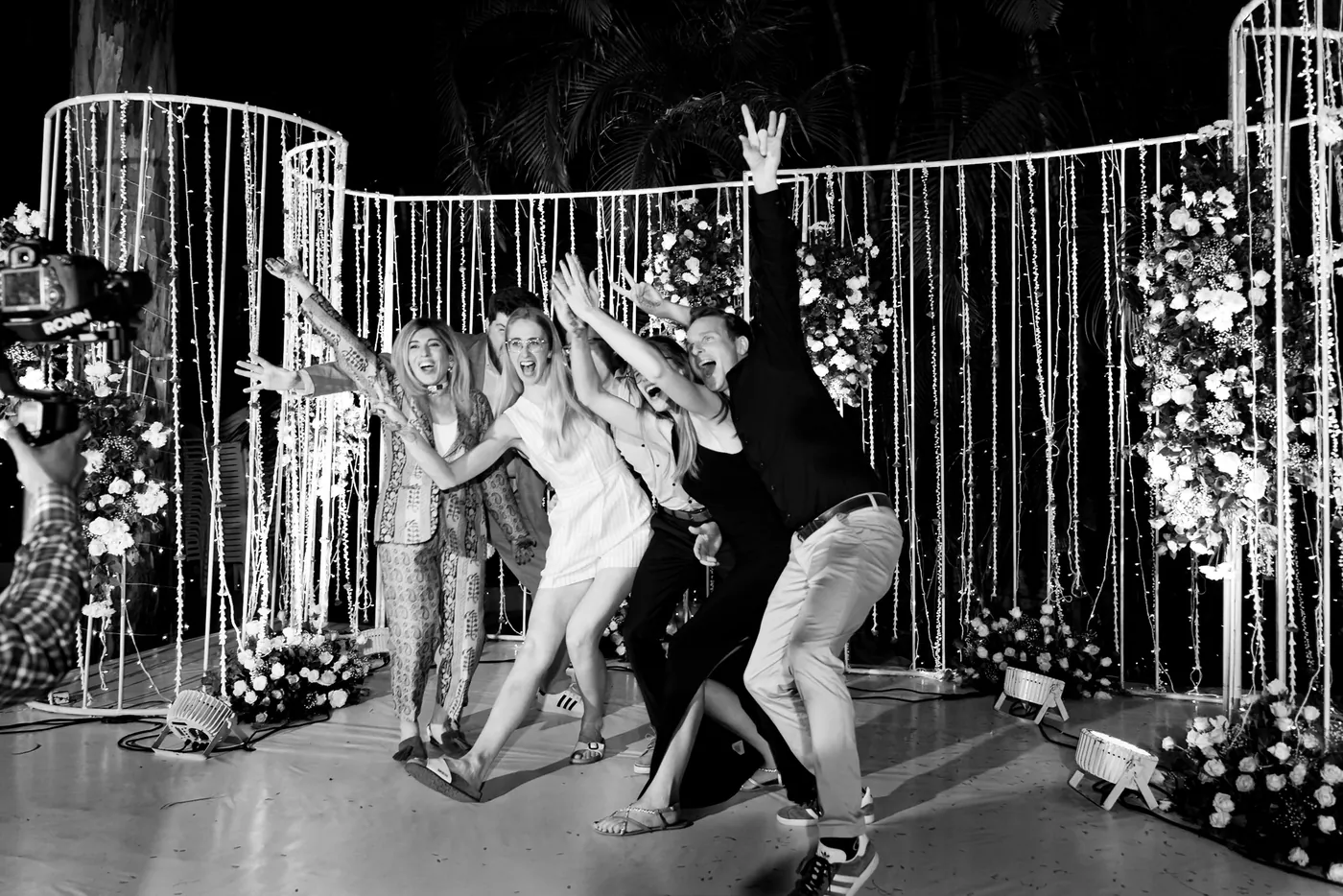 In black and white, a group dances with arms flung up in front of a curtain of lights