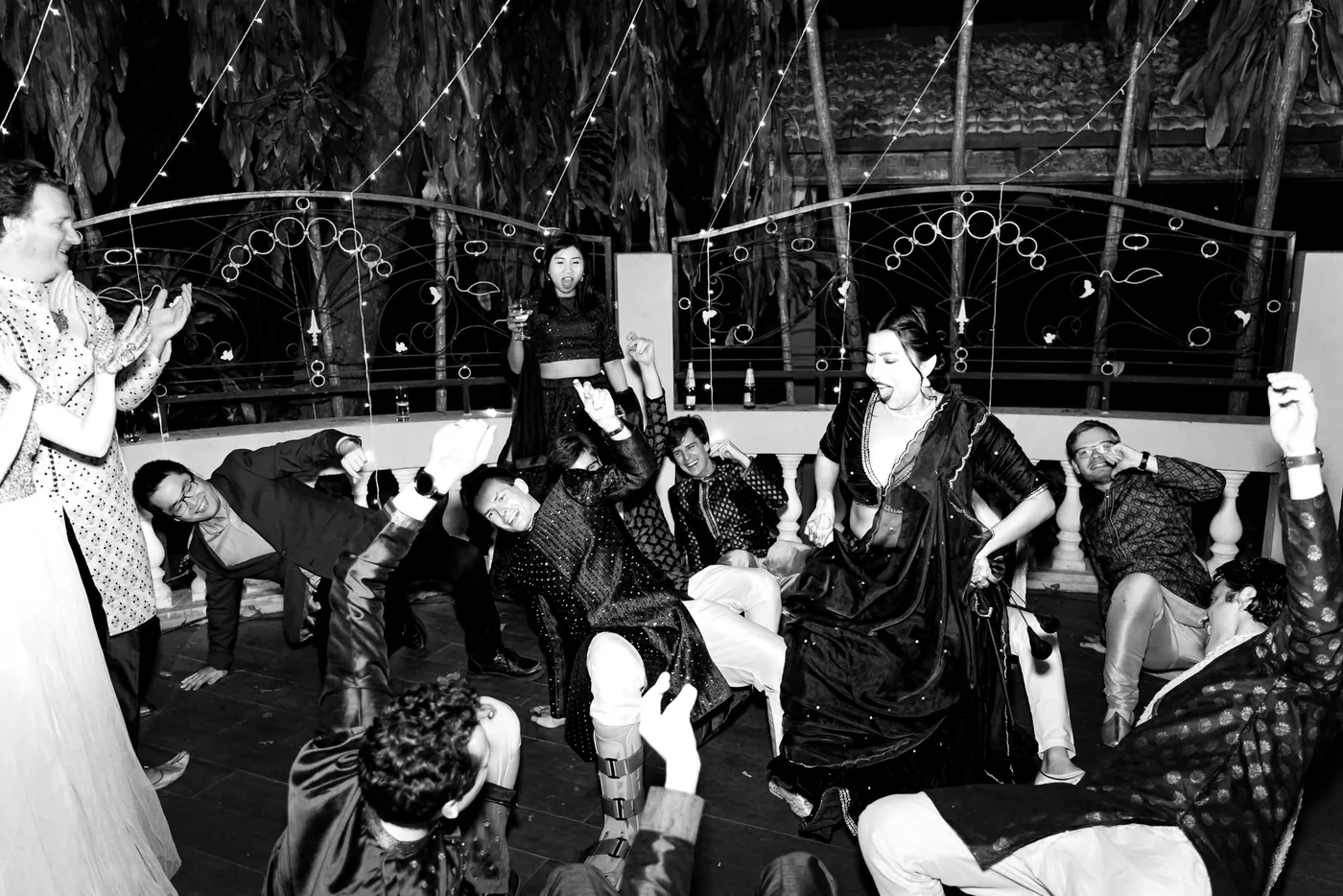 In black and white, guests sit low and clap around a woman in a dark dress dancing at the centre