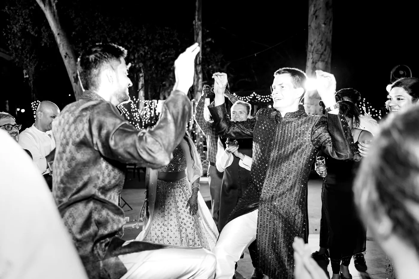 In black and white, two men in kurtas dance with raised hands as guests crowd close behind